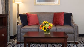 Habitación de hotel con sofá gris, dos almohadas rojas y una azul, una mesa de centro de madera, un jarrón con flores, un cuadro enmarcado en la pared y una alfombra estampada.