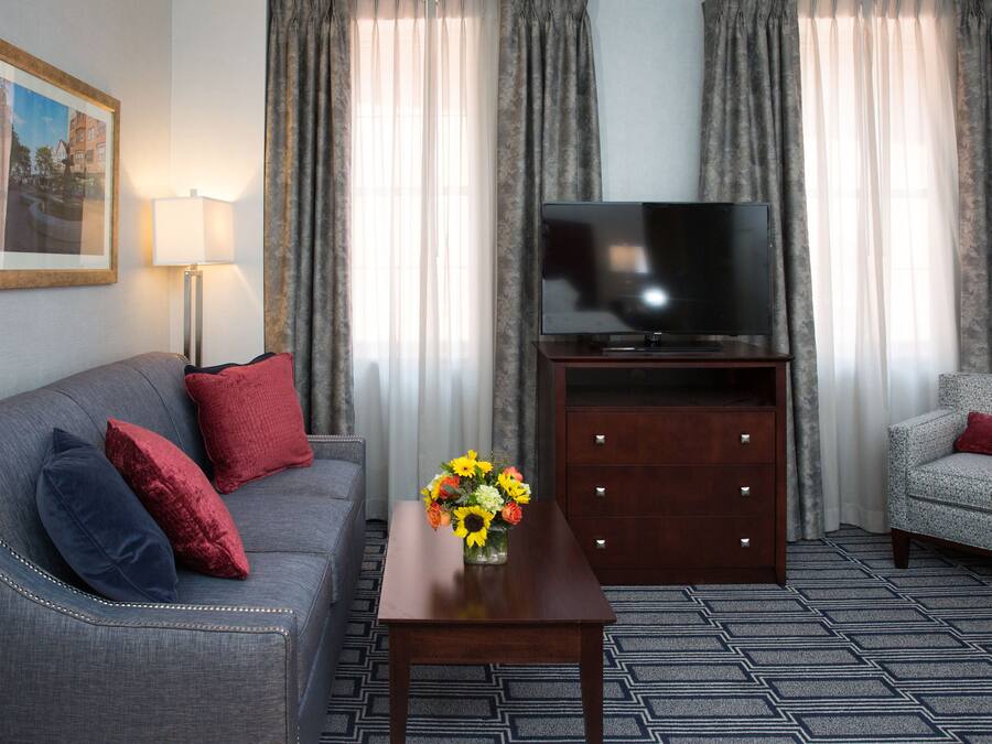 A living room with a grey sofa, red and blue cushions, a wooden coffee table with a vase of flowers, a flat-screen TV on a wooden stand, and patterned carpet.