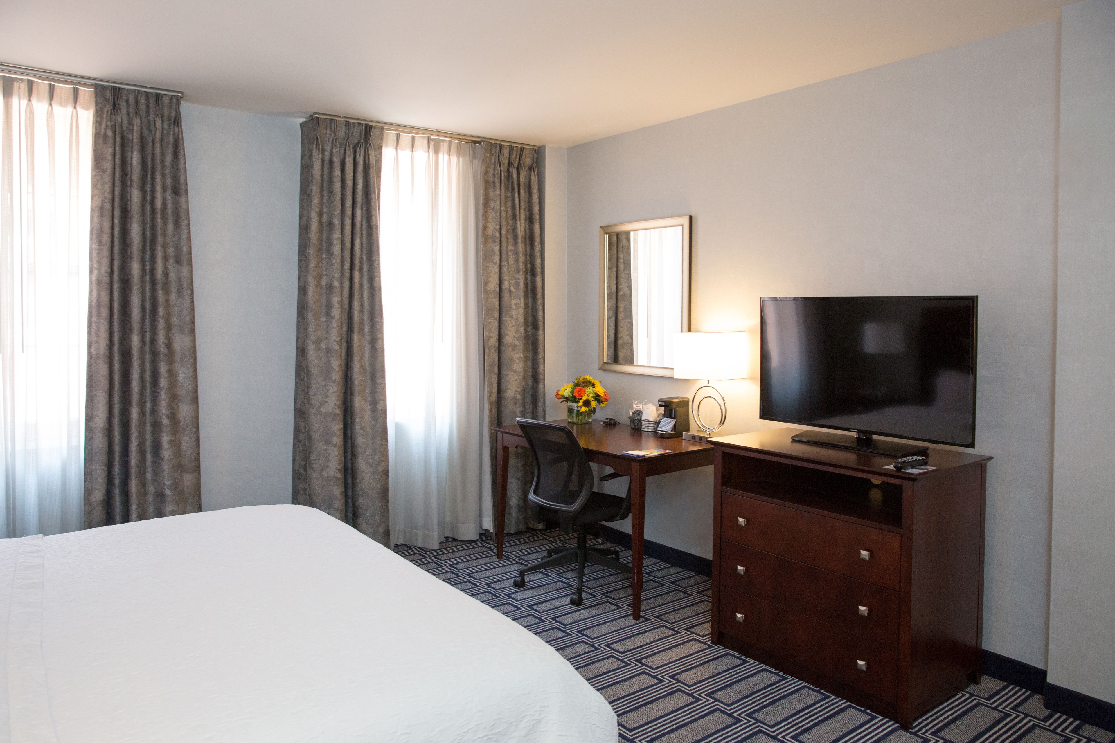 A hotel room with a large bed, a desk with a chair, a television, and a mirror.