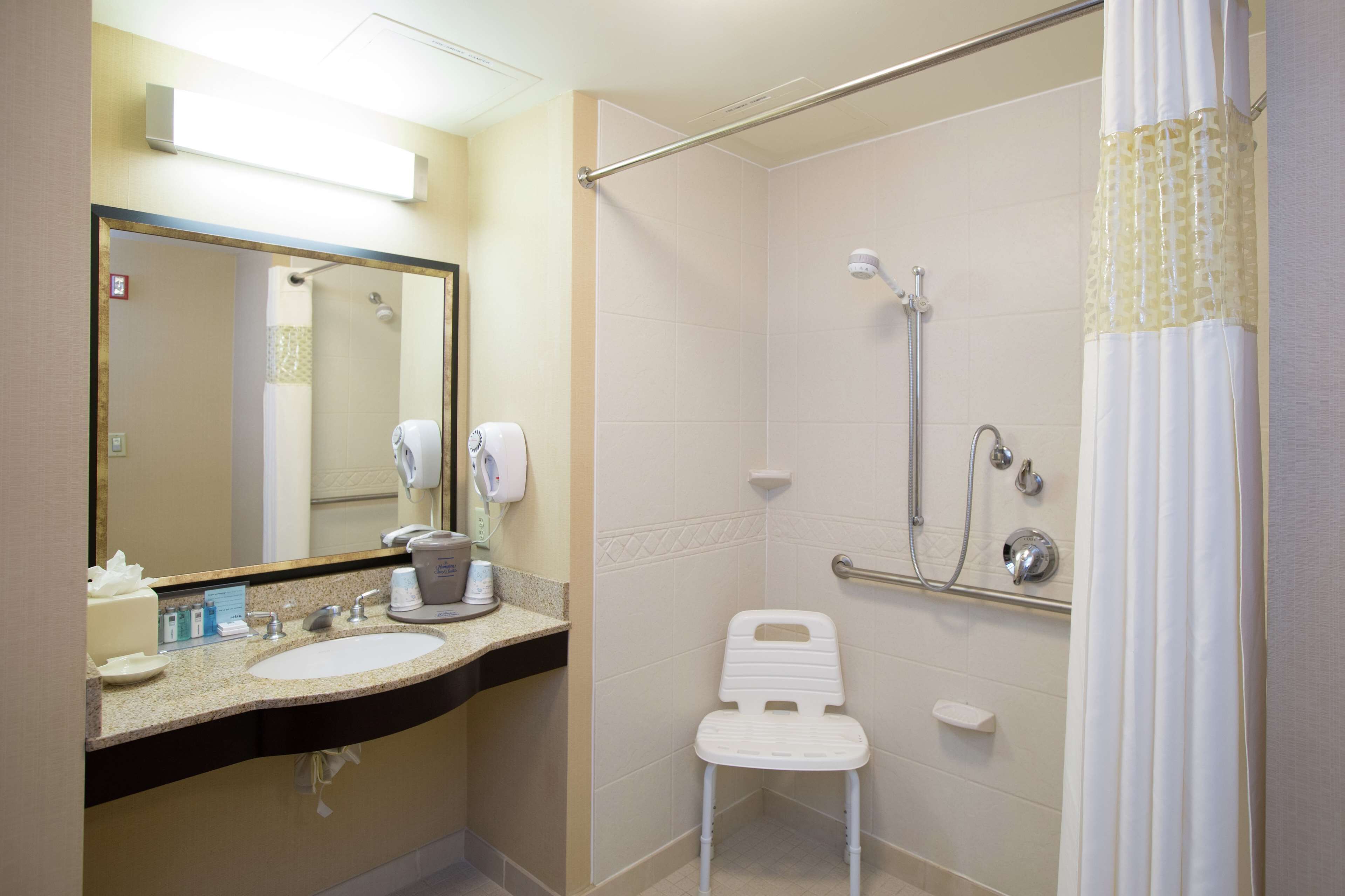 A bathroom with a sink, mirror, towel dispenser, and a shower with a grab bar.
