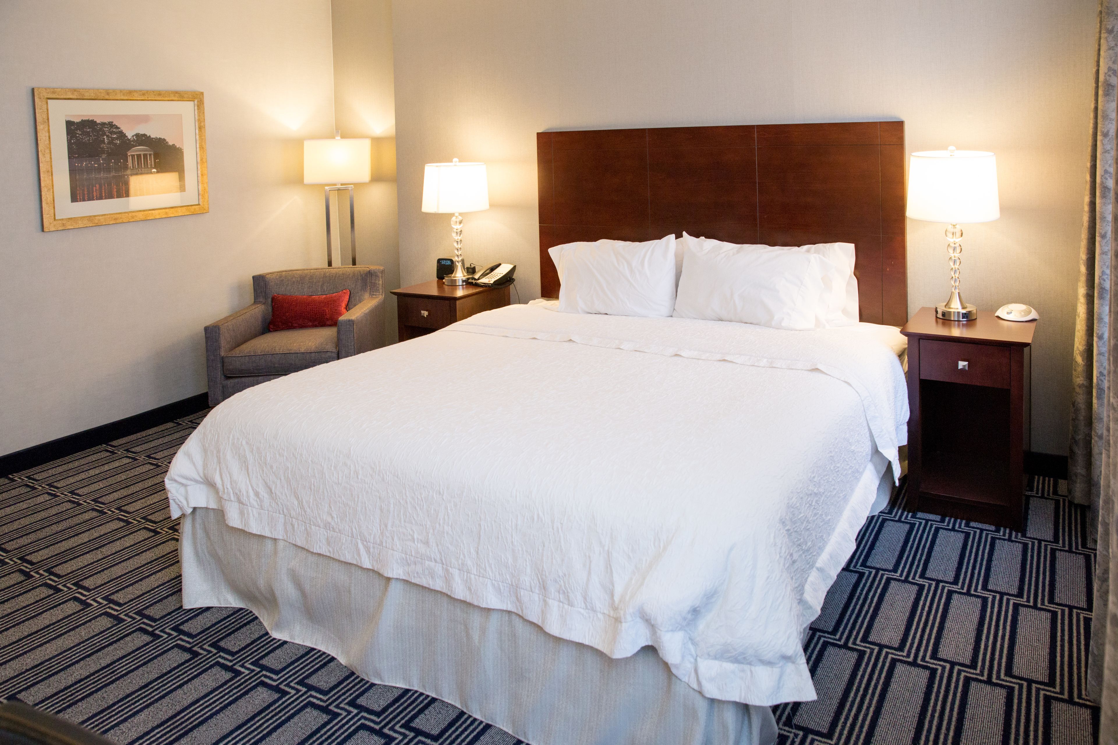 Habitación de hotel con una cama grande, dos mesitas de noche con lámparas, una silla e imagen enmarcada en la pared.