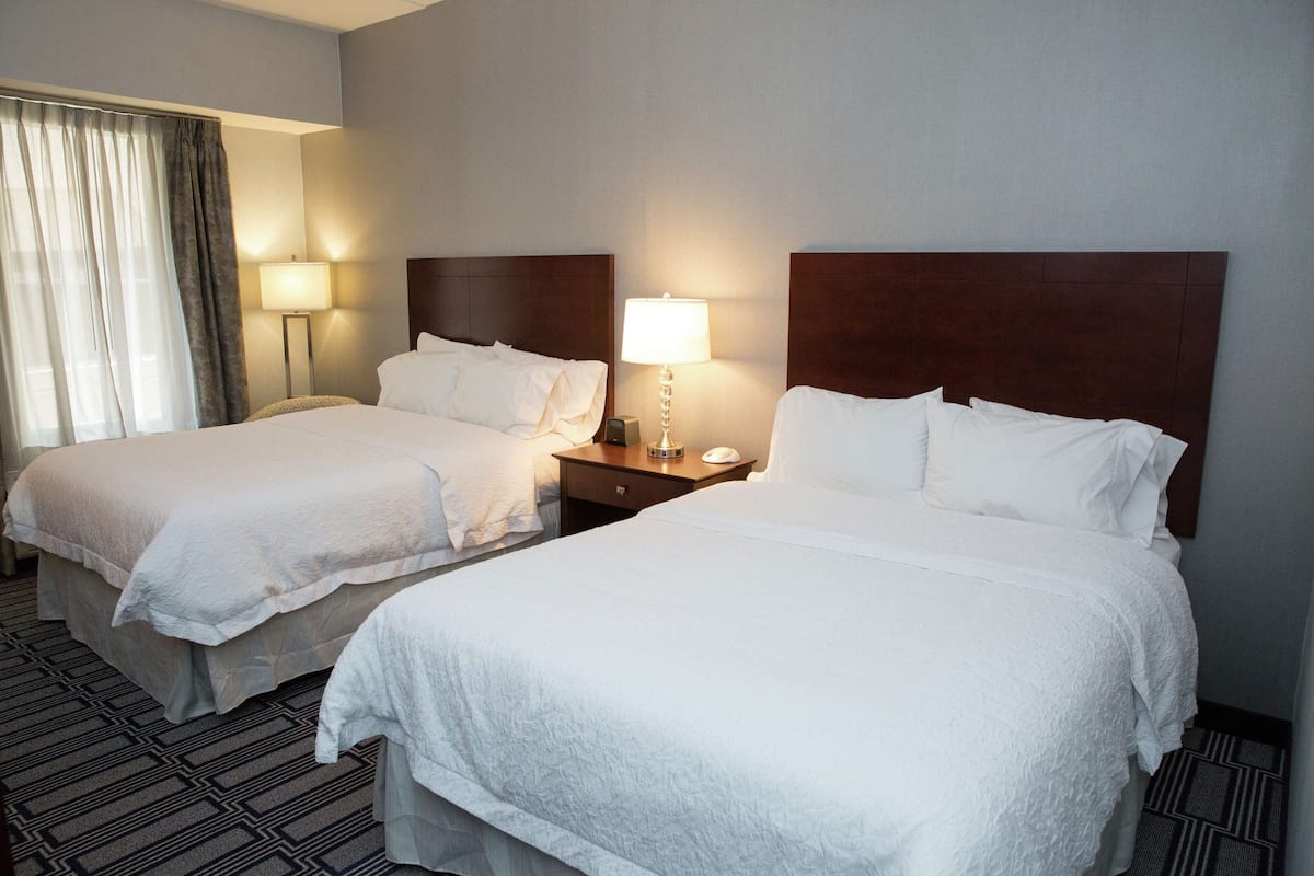 Une chambre d’hôtel avec deux lits, chacun équipé de draps blancs, une table de chevet avec une lampe et une fenêtre avec des rideaux.