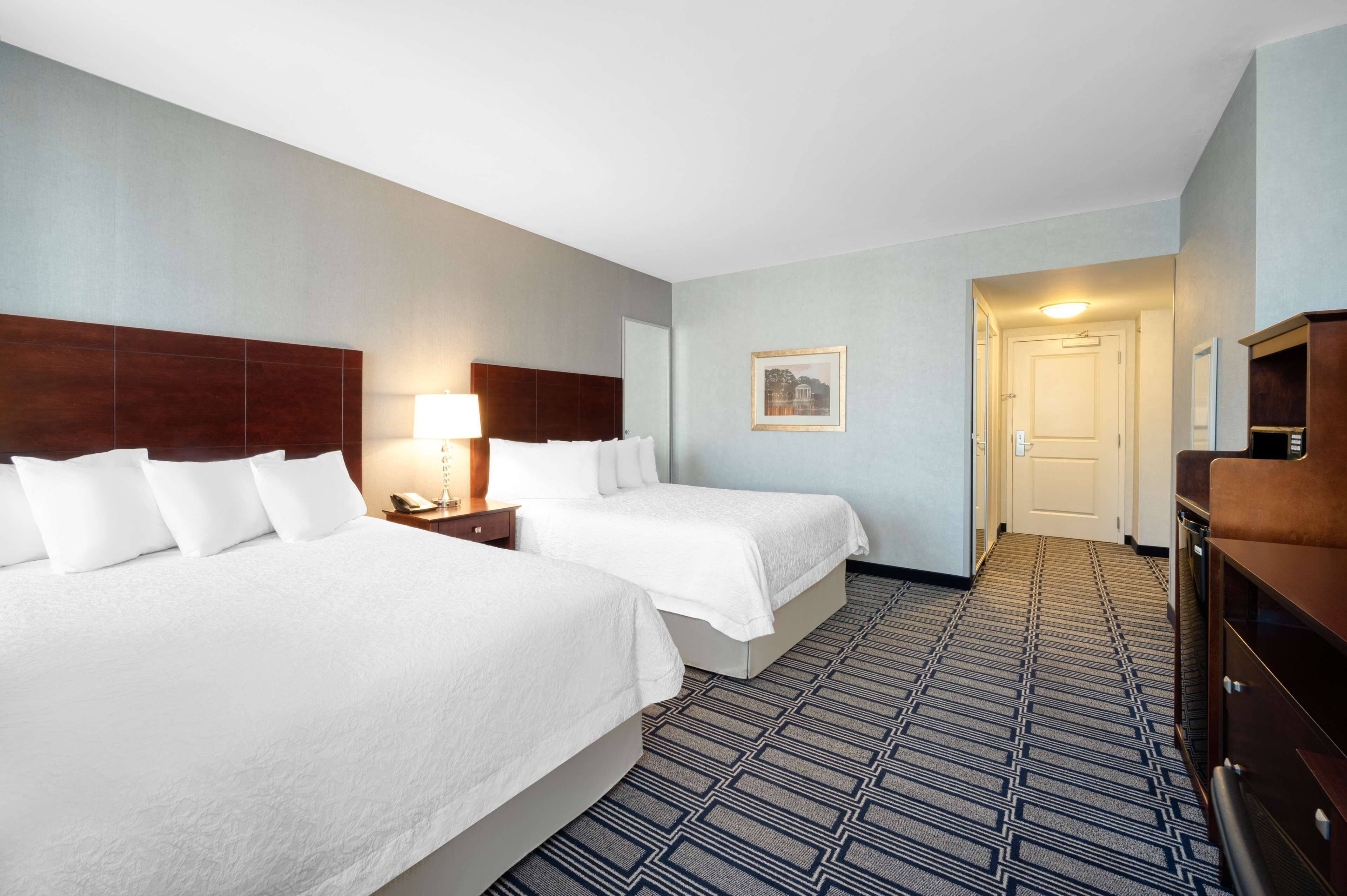 Quarto de hotel com duas camas, cabeceira de madeira, mesa de cabeceira com abajur, quadro na parede e cômoda.