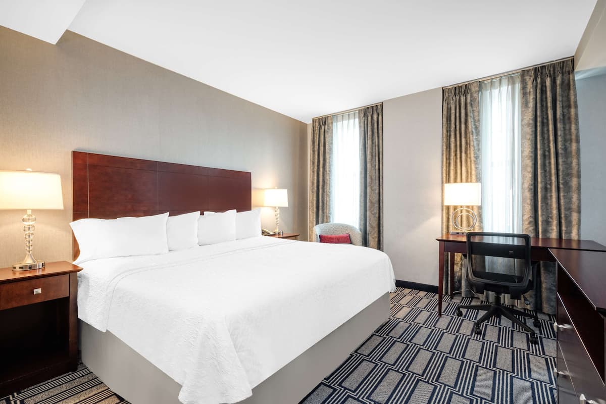 Une chambre d’hôtel avec un grand lit, deux tables de chevet avec des lampes, un bureau avec une chaise et une grande fenêtre avec des rideaux.