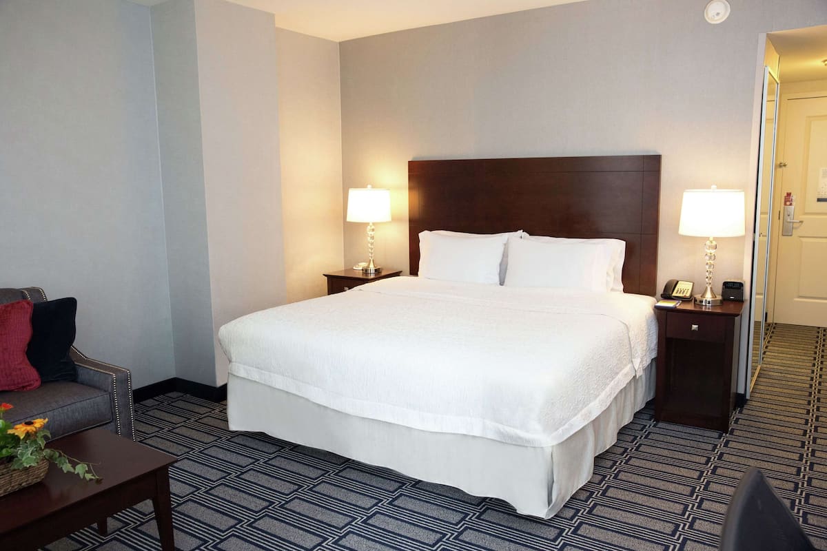 Une chambre d’hôtel avec un grand lit, deux tables de chevet avec des lampes, une petite table avec un panier, et un canapé avec un coussin rouge.