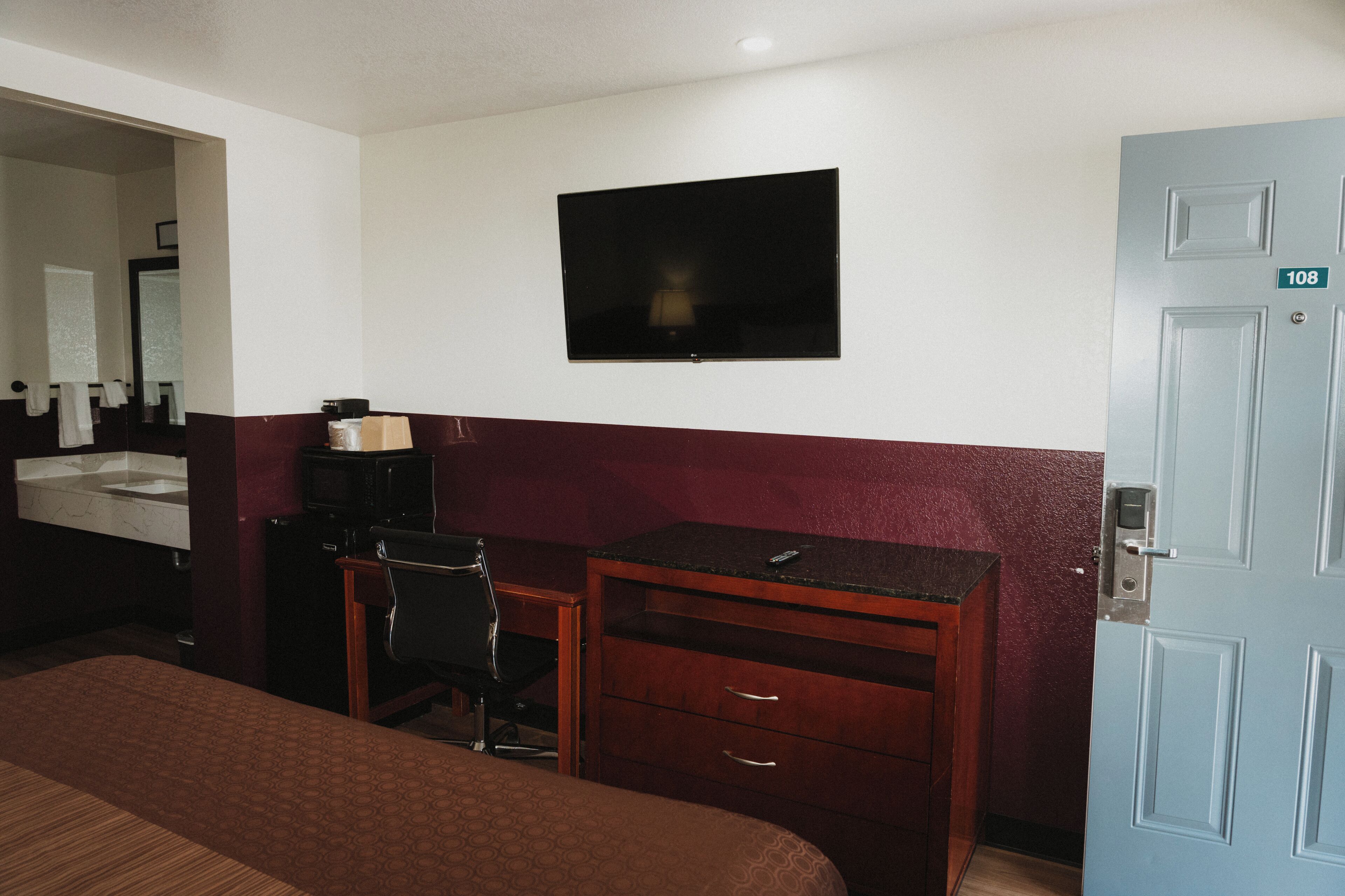 A hotel room with a bed, a desk with a chair, a television, a bathroom with a sink, and a door marked with the number 108.