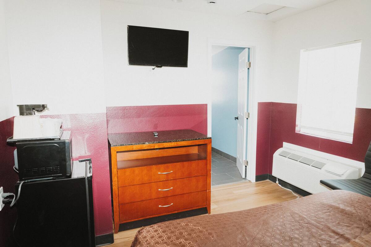 Habitación de hotel con cama, cómoda de madera, televisor de pantalla plana, calentador y puerta que da a otra habitación.