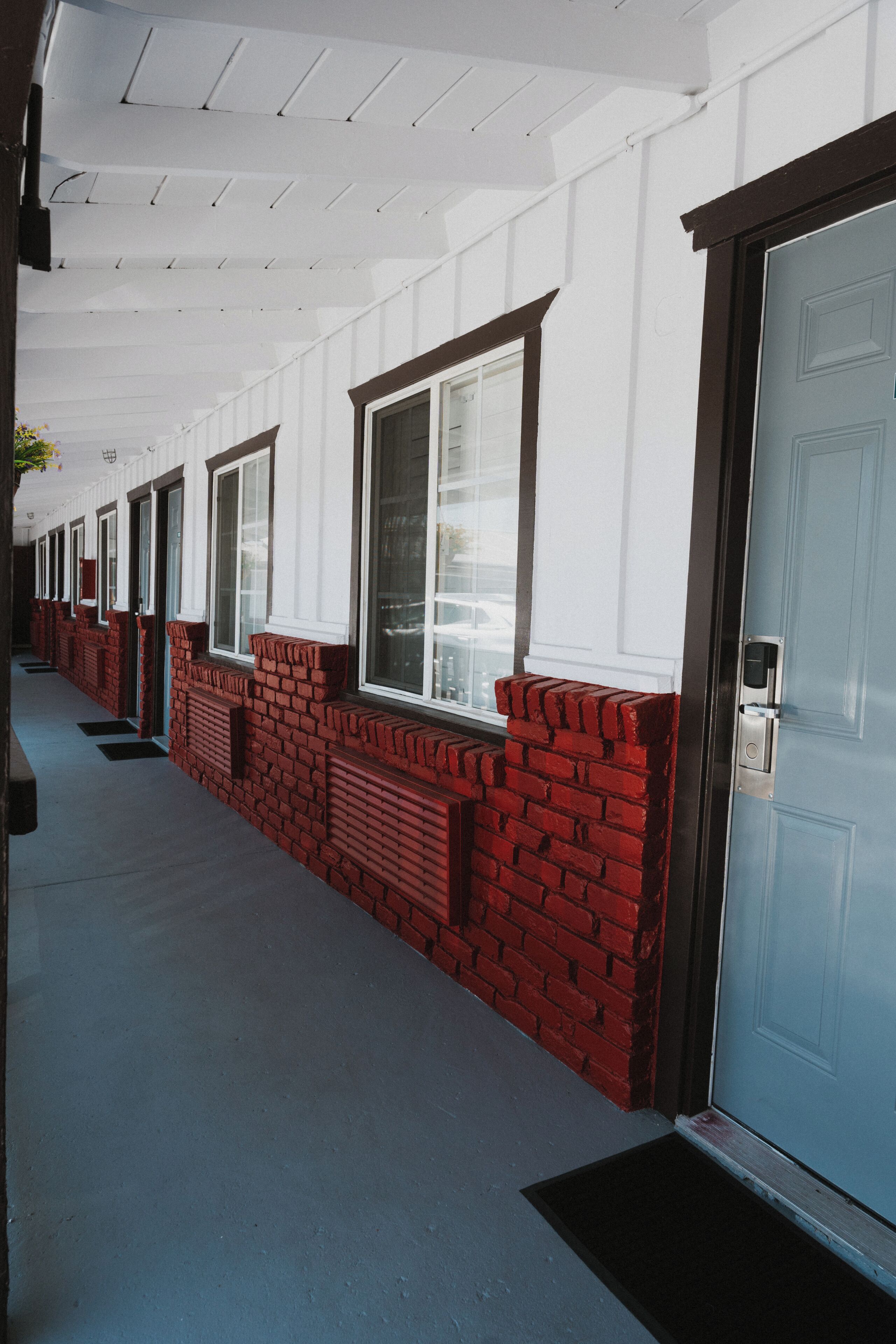 A row of hotel rooms with white walls and red brick accents, featuring large windows and a covered walkway.