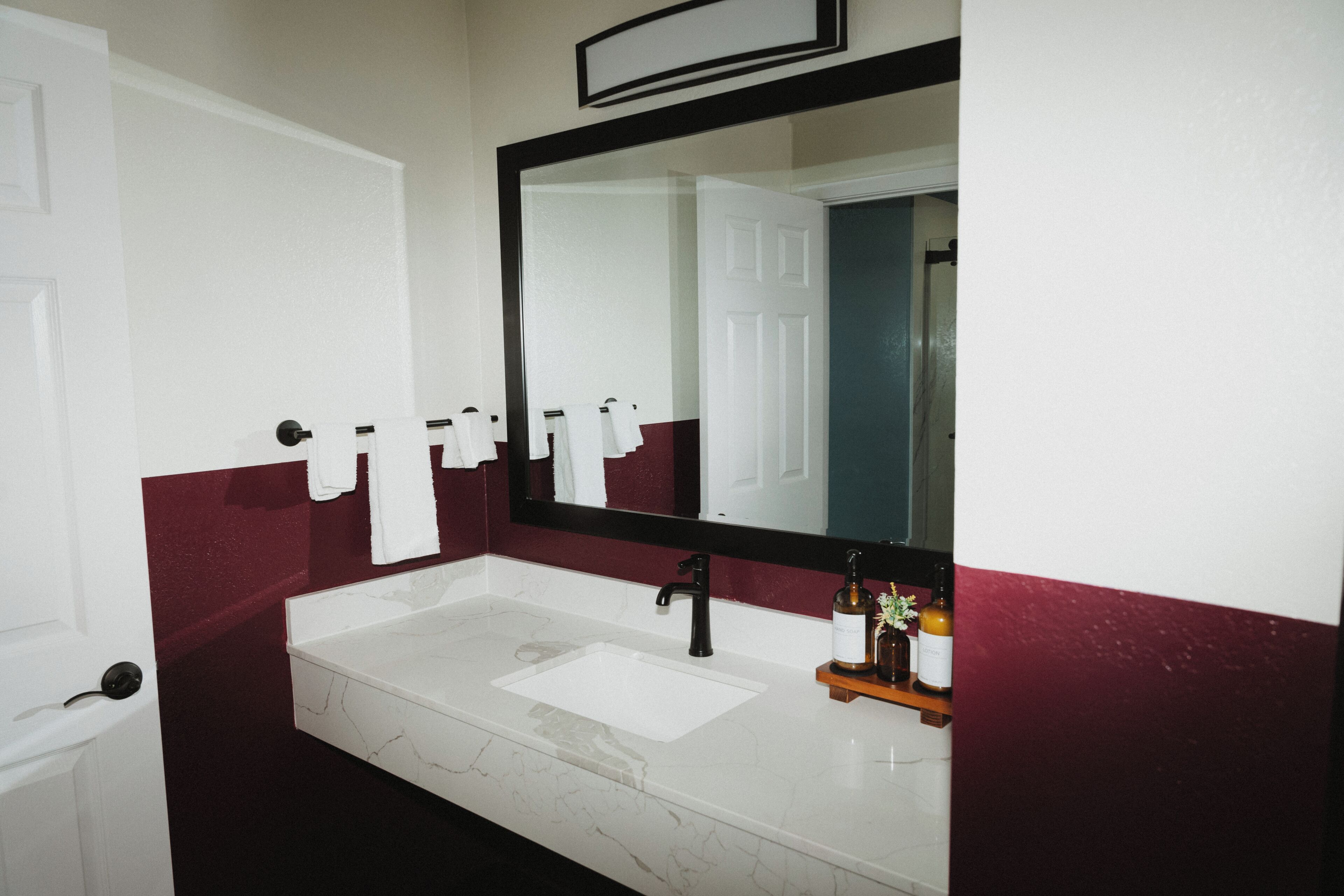 A bathroom with a white sink, black faucet, and a mirror above it. There are towels hanging on a rack and a bottle of shampoo on the counter.