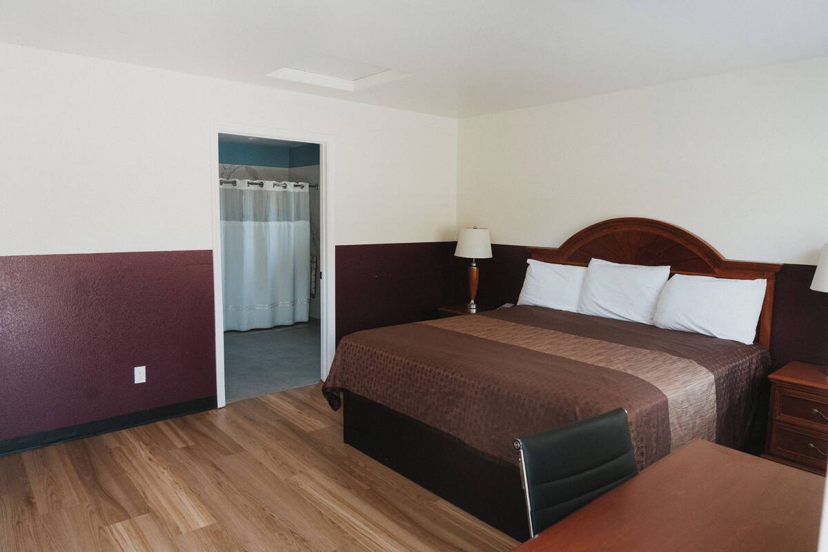 Una habitación de hotel con piso de madera, una cama con cubrecama marrón, una mesita de noche y una lámpara.