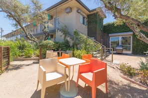 Un espace extérieur aménagé avec des tables et des chaises, un escalier menant à un bâtiment, et un jardin avec des plantes et des arbres.