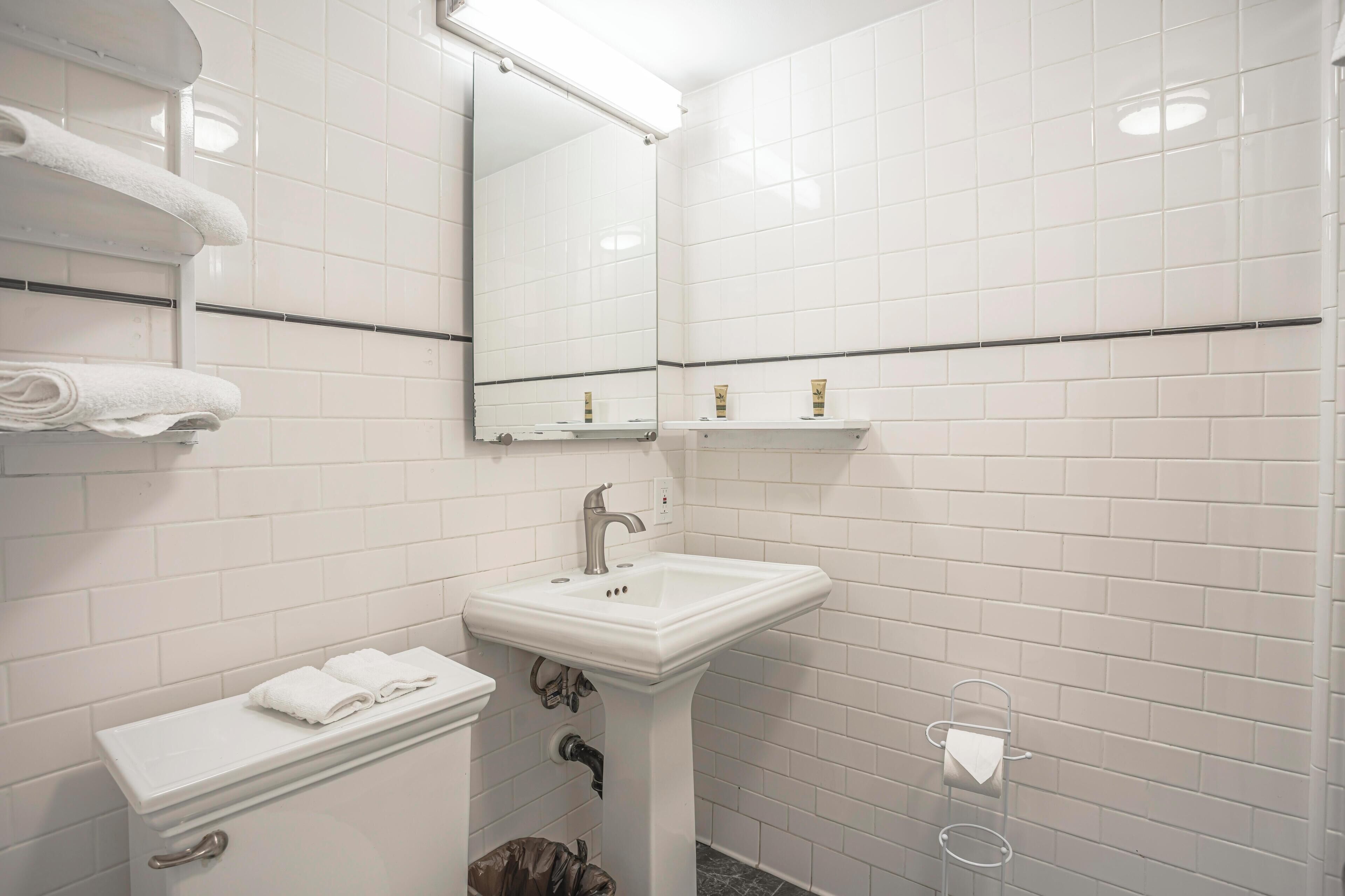 Un baño con paredes de azulejos blancos, un lavamanos pedestal, un espejo, un perchero para toallas y un inodoro.