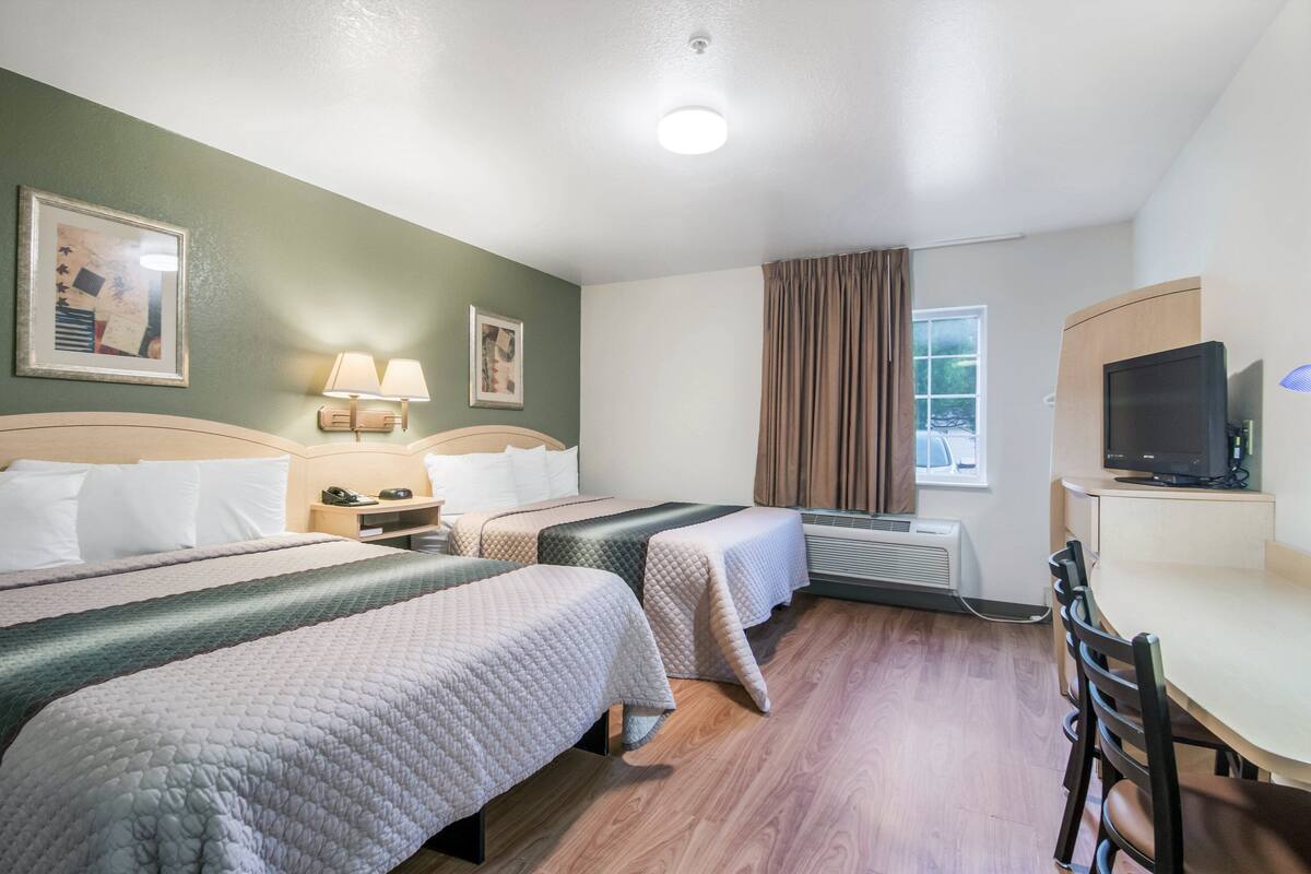 Ein Hotelzimmer mit zwei Betten, einem Schreibtisch, einem Stuhl, einem Fernseher und einem Vorhangfenster.