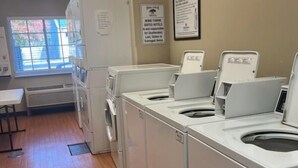 A laundry room with four washing machines and dryers, a white table, and a window with blinds. - HomeTowne Studios & Suites by Red Roof Bentonville (Bentonville)