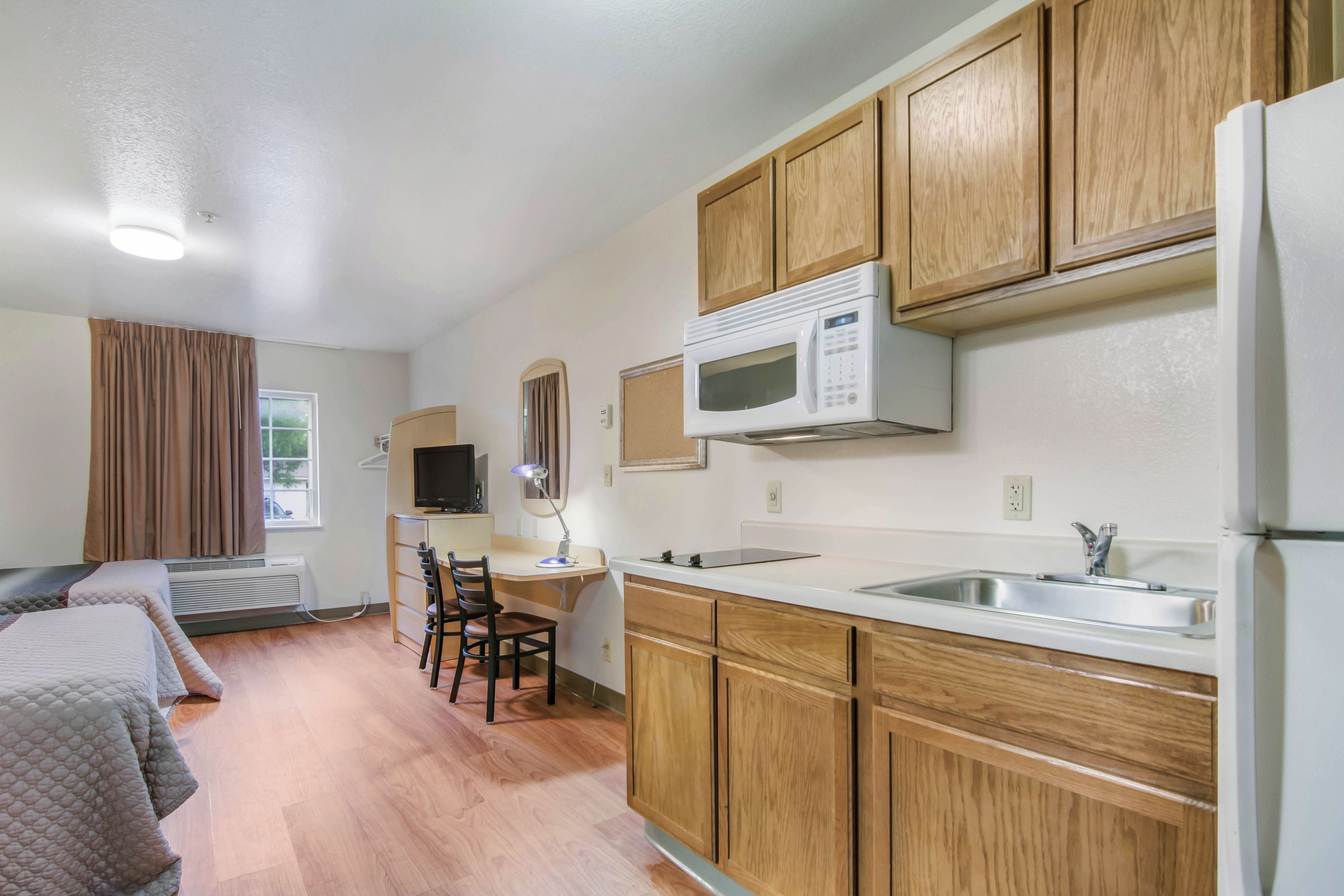 A compact kitchenette with wooden cabinets, a microwave, a sink, and a small desk with a chair.