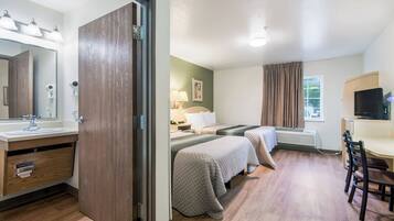Habitación de hotel con piso de madera, cama con colcha, escritorio con silla, televisor y baño con espejo y lavamanos.