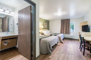 Habitación de hotel con piso de madera, cama con colcha, escritorio con silla, televisor y baño con espejo y lavamanos.