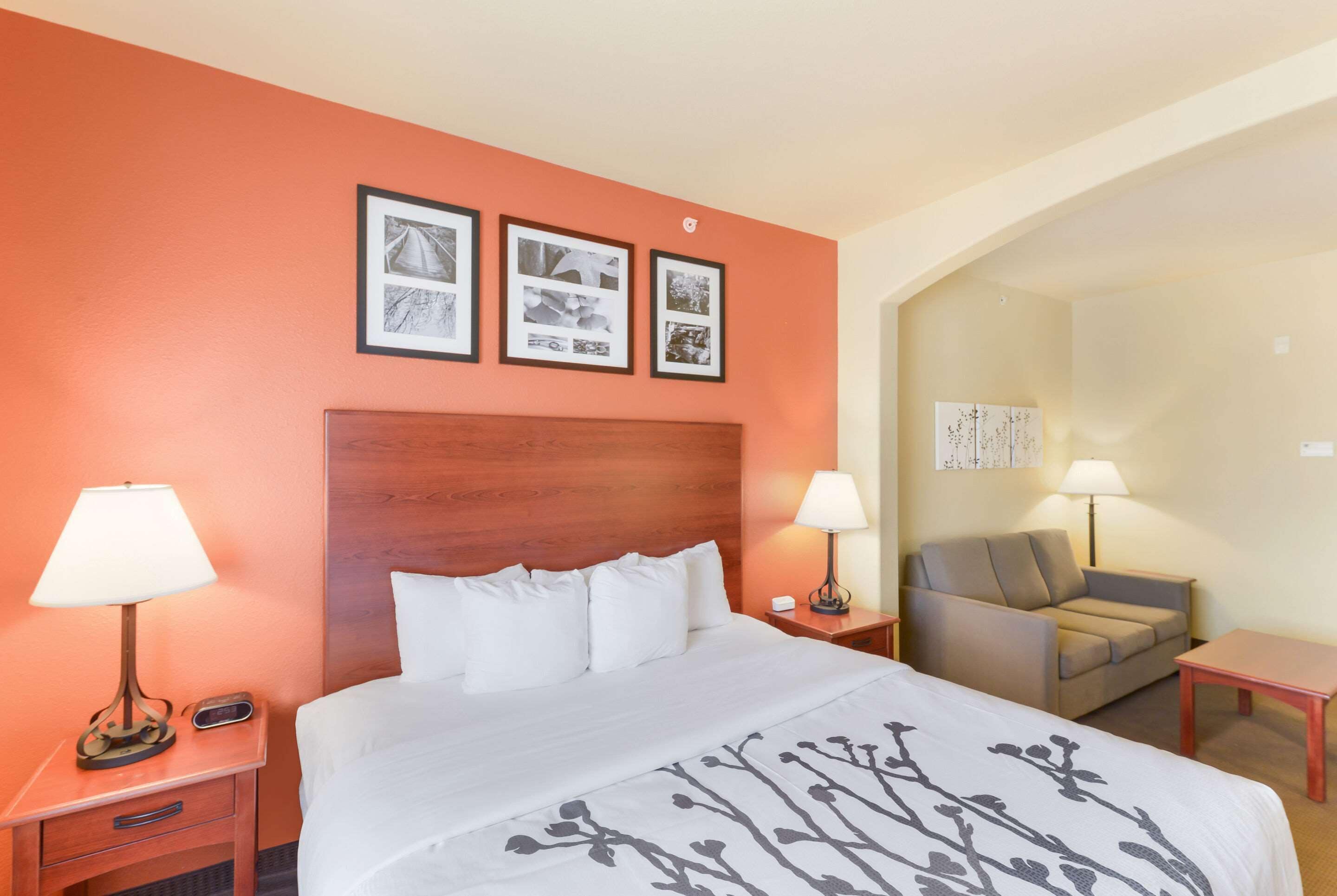 A hotel room with a bed, two bedside tables, a chair, and a small table. There are framed pictures on the wall above the bed.