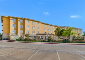 A multi-story hotel with a yellow facade, a parking lot, and a landscaped area with trees and shrubs.