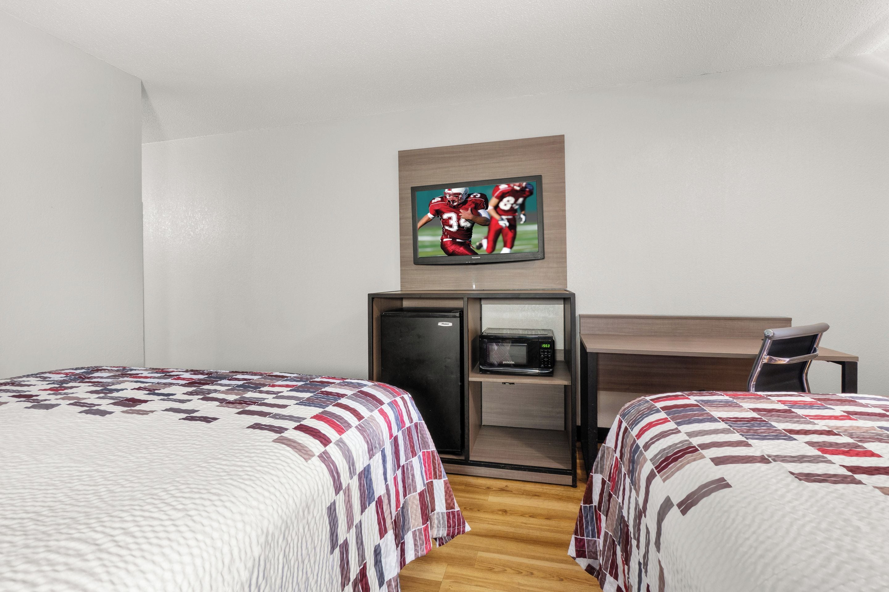 A hotel room with two beds, a desk, a refrigerator, and a television on the wall.