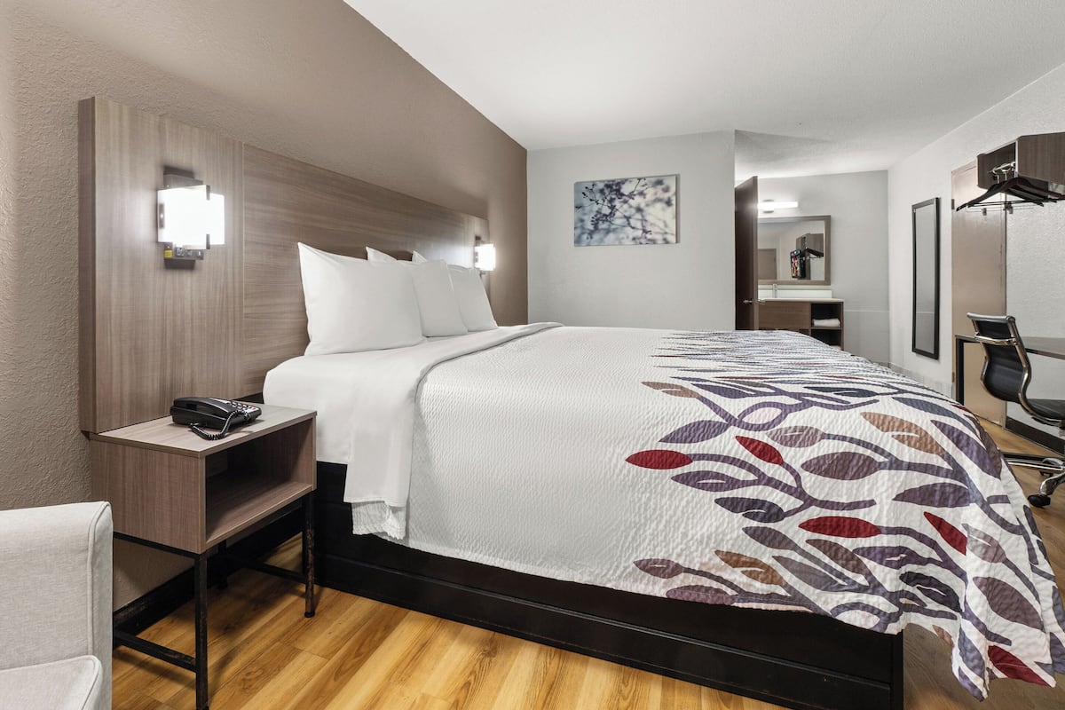 Ein Hotelzimmer mit einem großen Bett, einem Nachttisch mit Telefon, einem Sessel, einem Schreibtisch mit Stuhl und einem Spiegel.