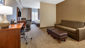 A hotel room with a wooden desk, a lamp, a sofa, an ottoman, a chair, a bed, and a TV. - Comfort Inn Louisville Southwest (Louisville)