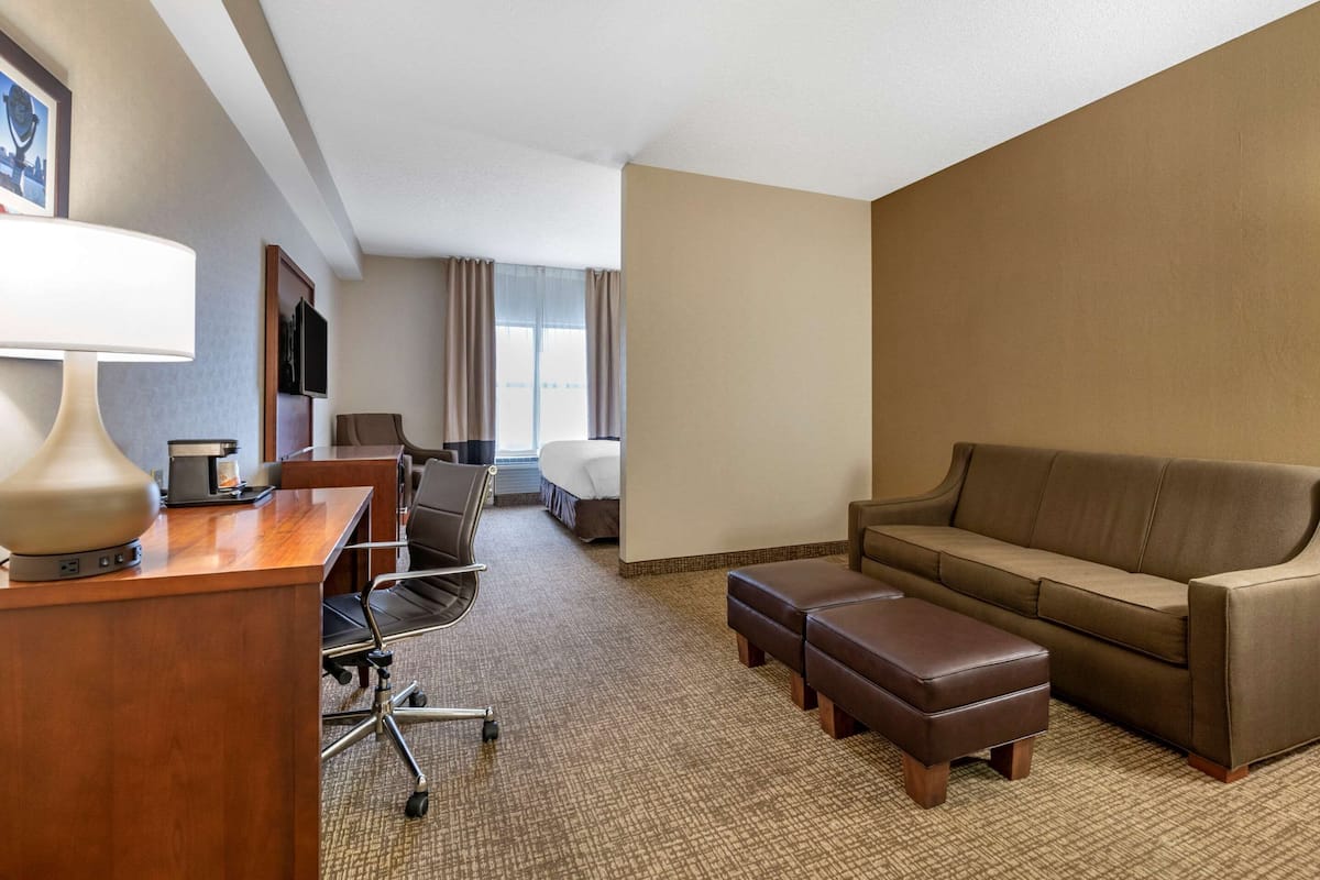 A hotel room with a wooden desk, a lamp, a sofa, an ottoman, a chair, a bed, and a TV.