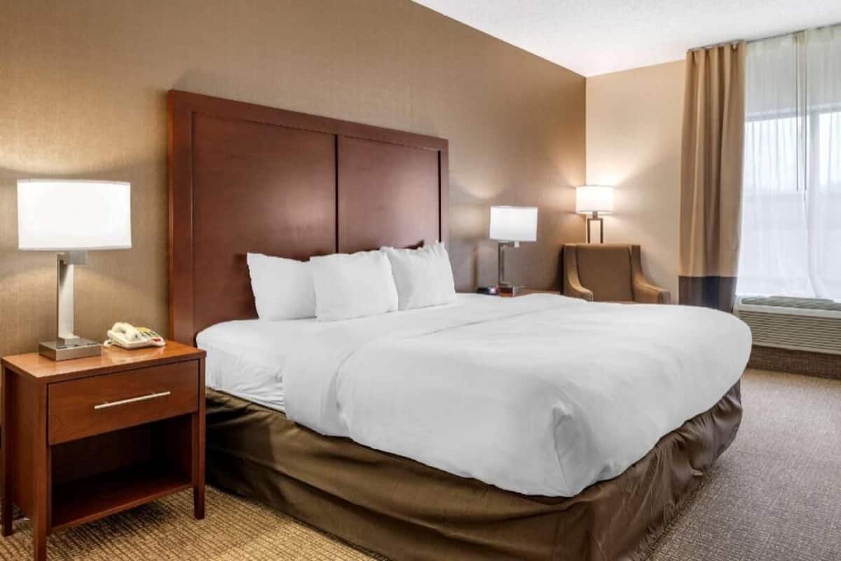 A hotel room with a large bed, a bedside table with a lamp, a chair, and a window with curtains.