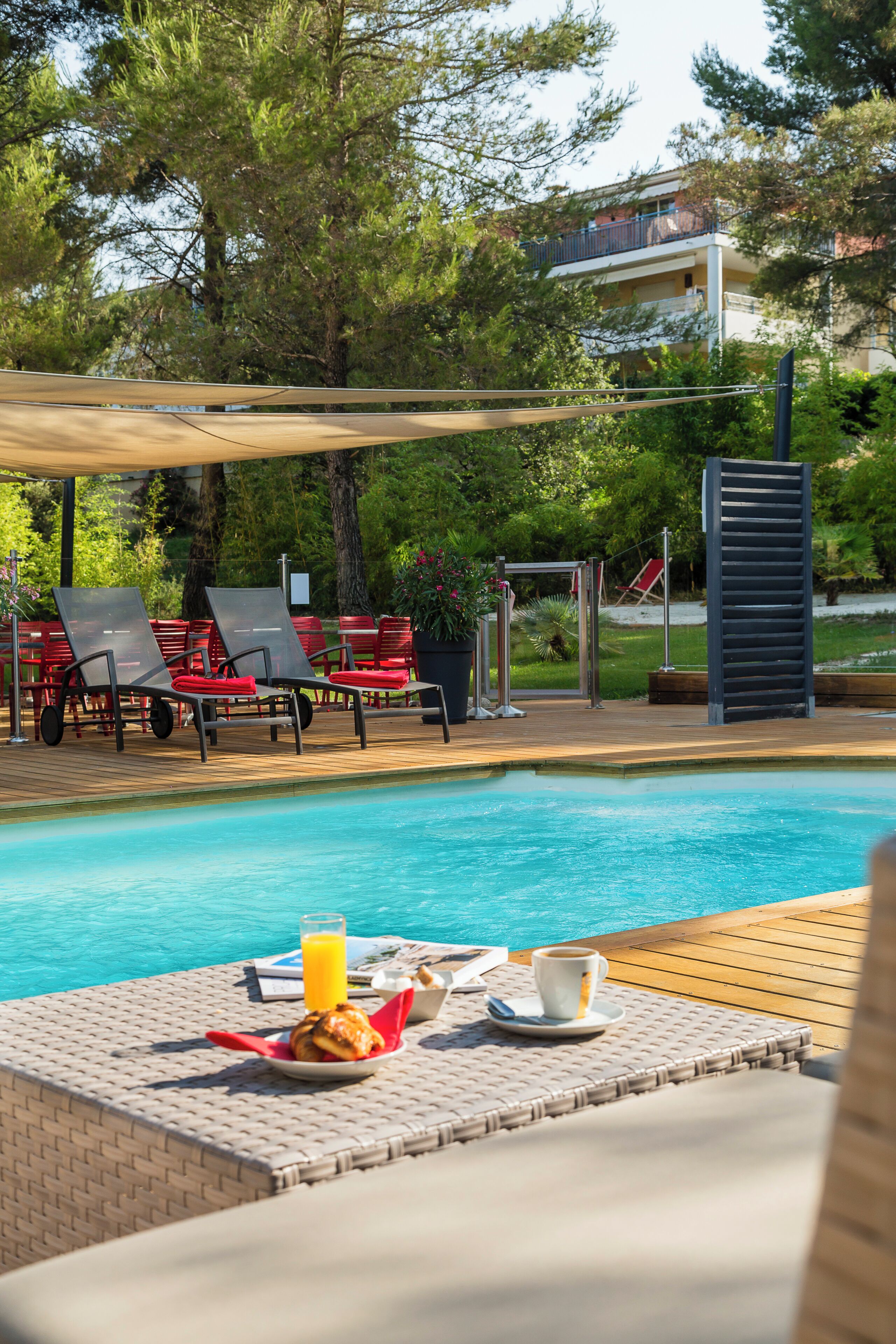 Un espace aménagé au bord de la piscine, avec des chaises longues, une table où sont disposés des éléments pour le petit-déjeuner, et une vue sur un bâtiment en arrière-plan.