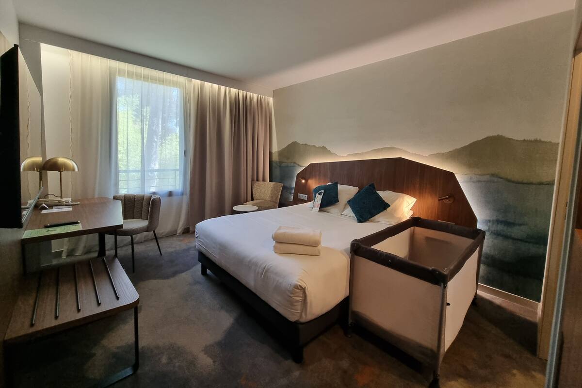 A hotel room with a bed, bedside table, chair, desk, and a large window with curtains.