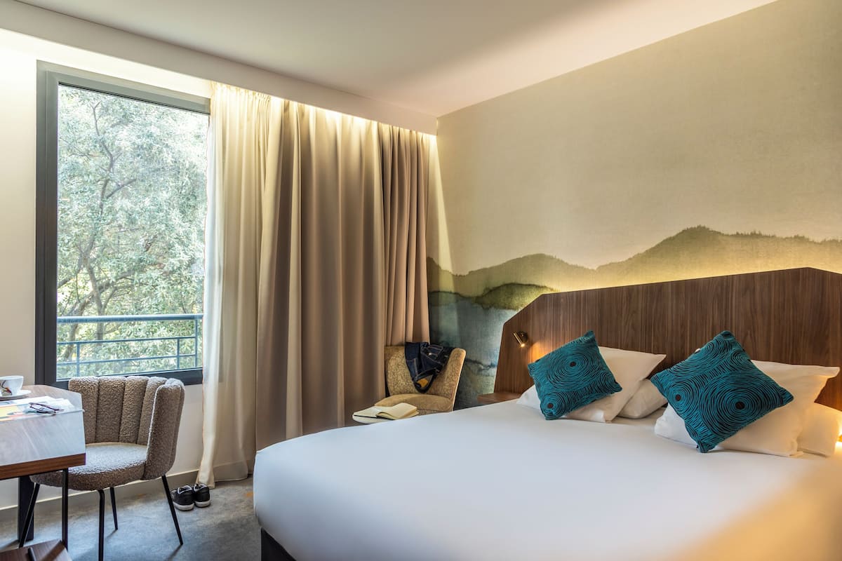 A hotel room with a large bed, a chair, a table, and a view of trees through a window.