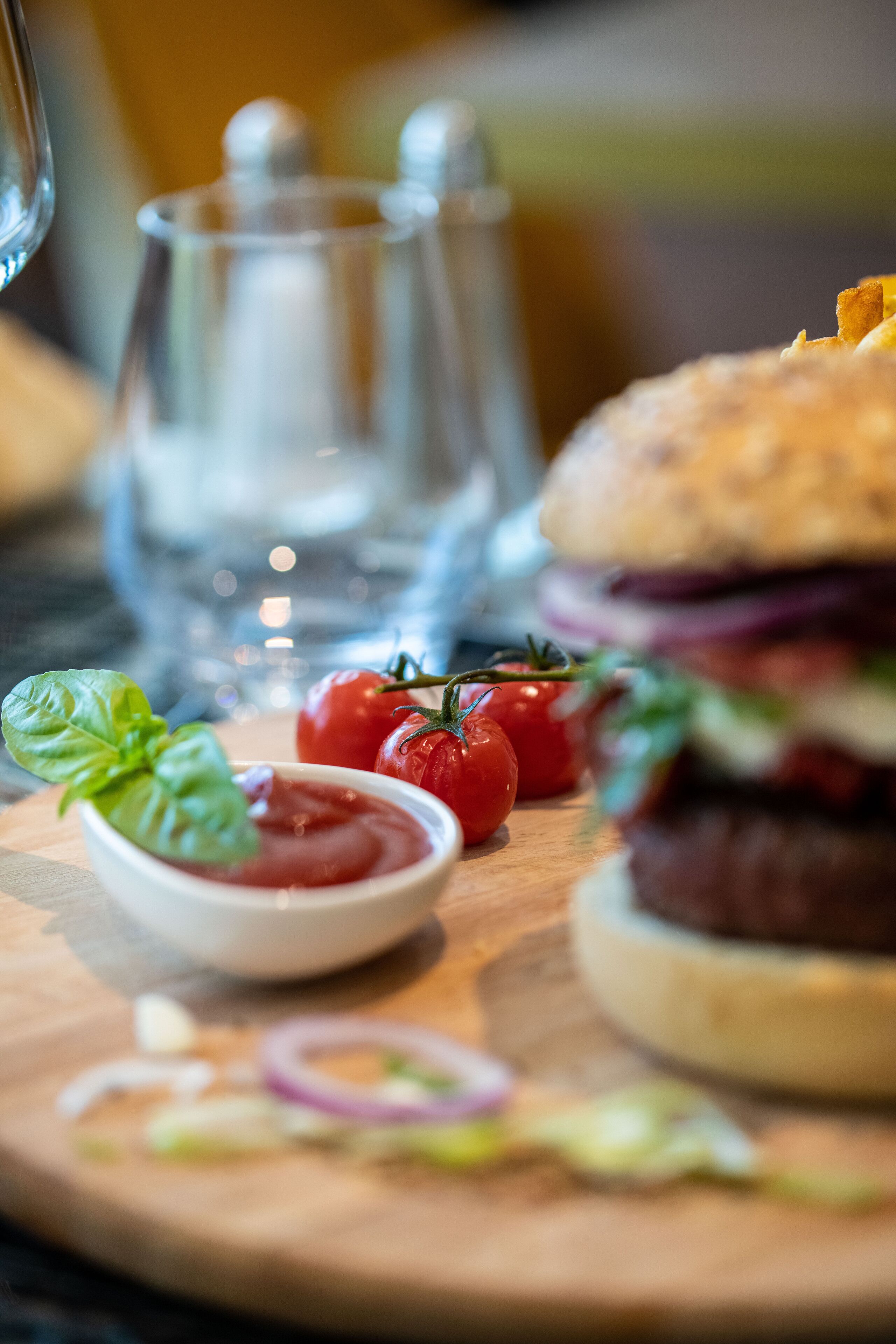 Una tabla de madera con una hamburguesa, tomates, albahaca y un recipiente pequeño de kétchup.