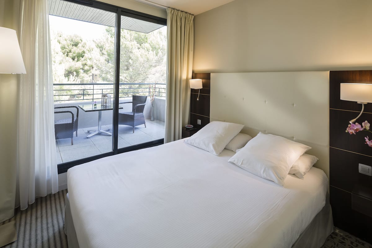 A hotel room with a large bed, a balcony with a table and chairs, and a view of trees.