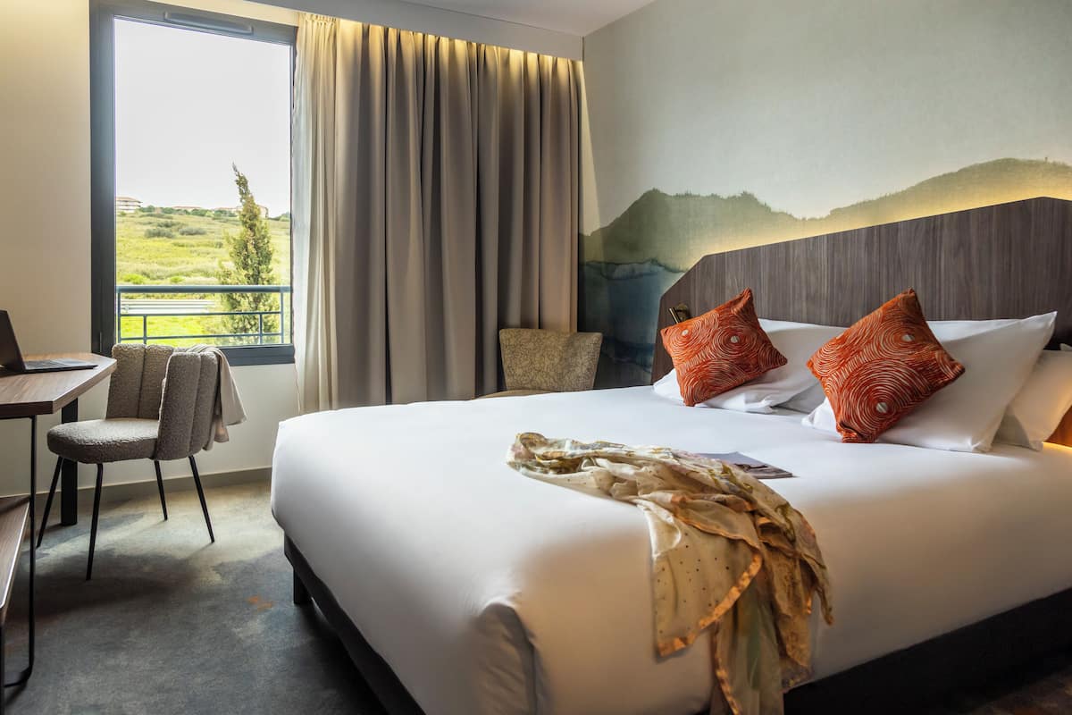 A hotel room with a large bed, a desk, and a chair. There is a window with curtains, a painting on the wall, and a view of a landscape.