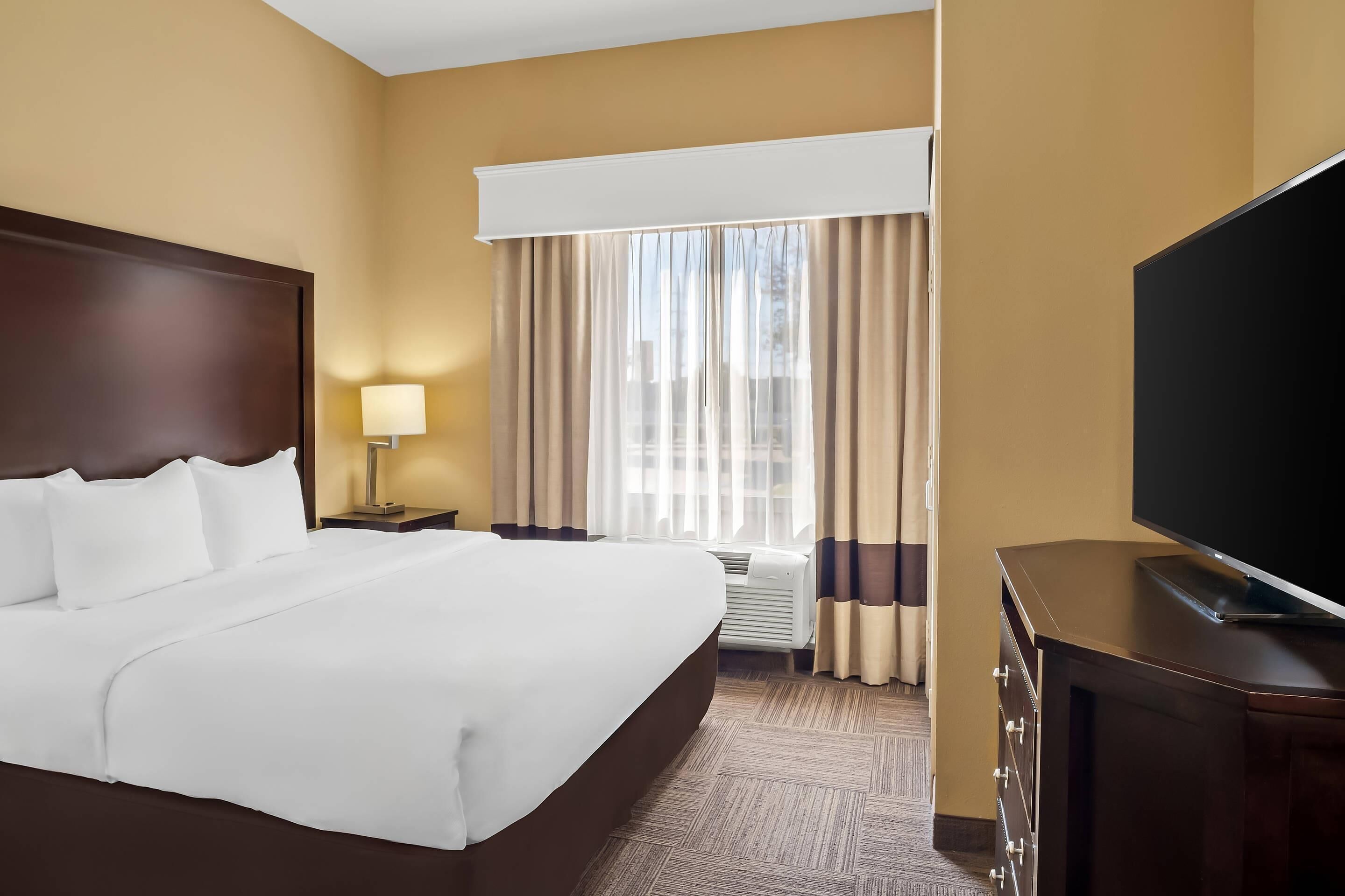 Habitación de hotel con una cama grande, un televisor de pantalla plana, una mesita de noche con lámpara y una ventana con cortinas.