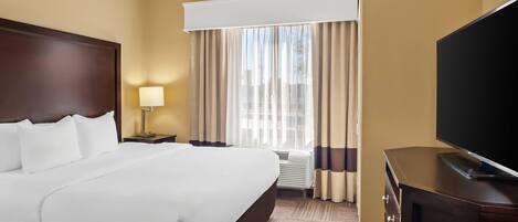 A hotel room with a large bed, a flat-screen TV, a nightstand with a lamp, and a window with curtains.