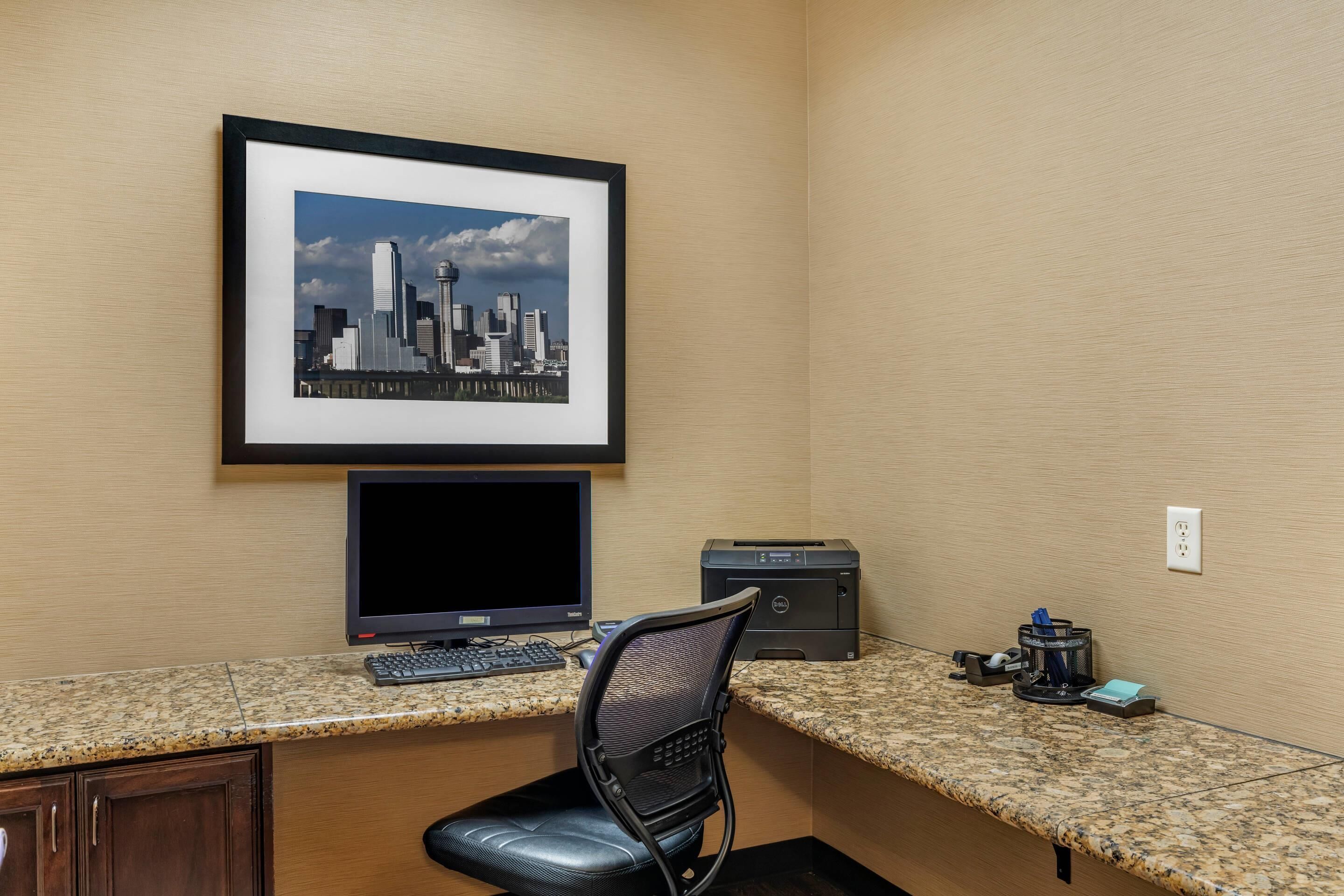 Un espace de travail comprenant un ordinateur de bureau, une imprimante, un tableau représentant un paysage urbain et une chaise.