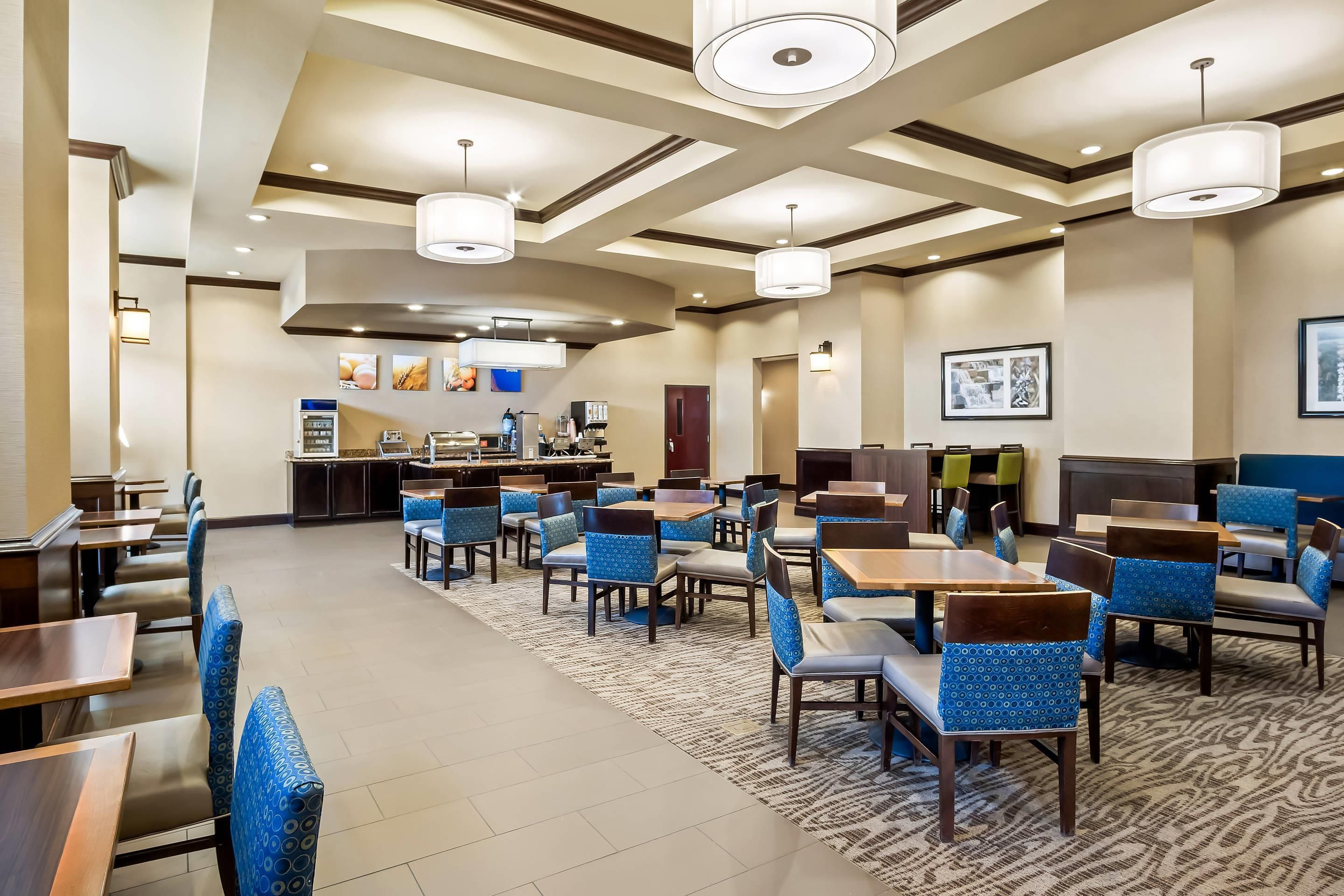 Uma sala de jantar de hotel com várias mesas e cadeiras, uma cozinha ao fundo e um tapete estampado.