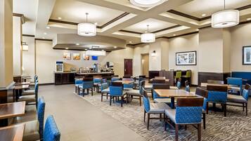 A hotel dining area with multiple tables and chairs, a kitchen in the background, and a patterned carpet.
