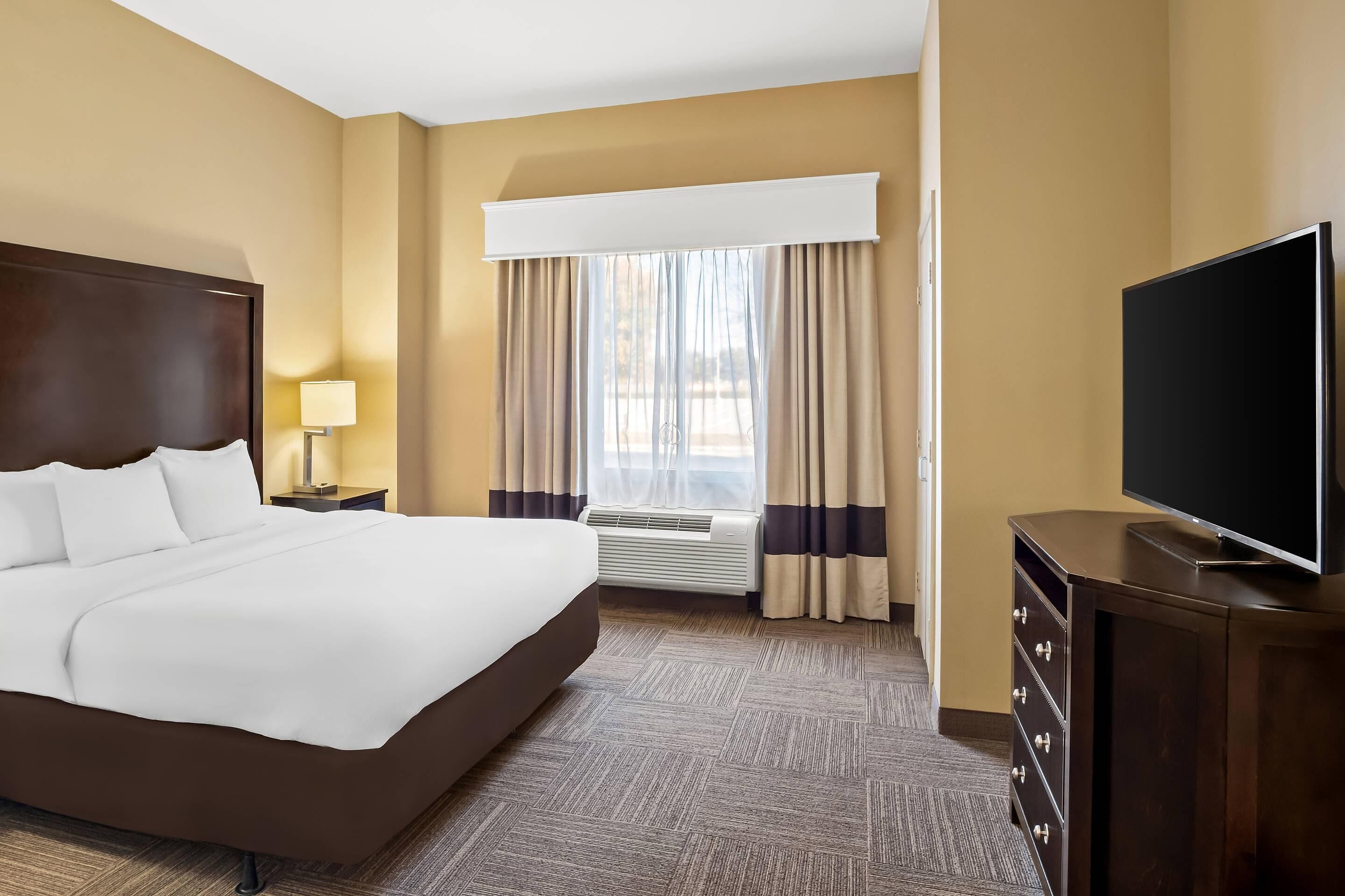 A hotel room with a large bed, a flat-screen TV, a dresser, a lamp, and a window with curtains.