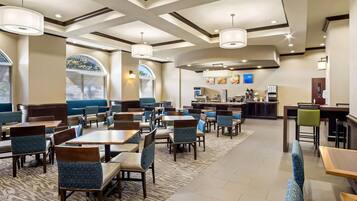 A hotel dining area with tables and chairs, a bar, and a view of the outdoors through large windows.