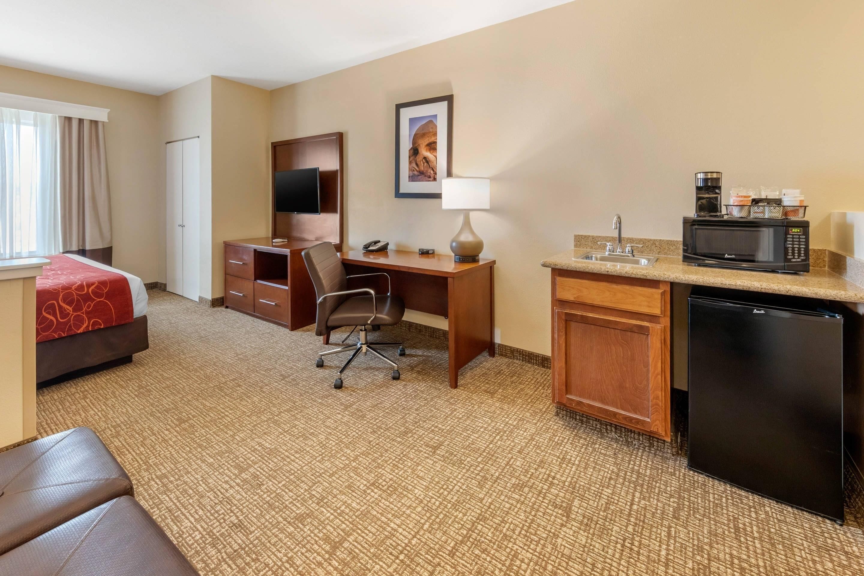 A hotel room with a bed, desk, chair, kitchenette, and a TV.