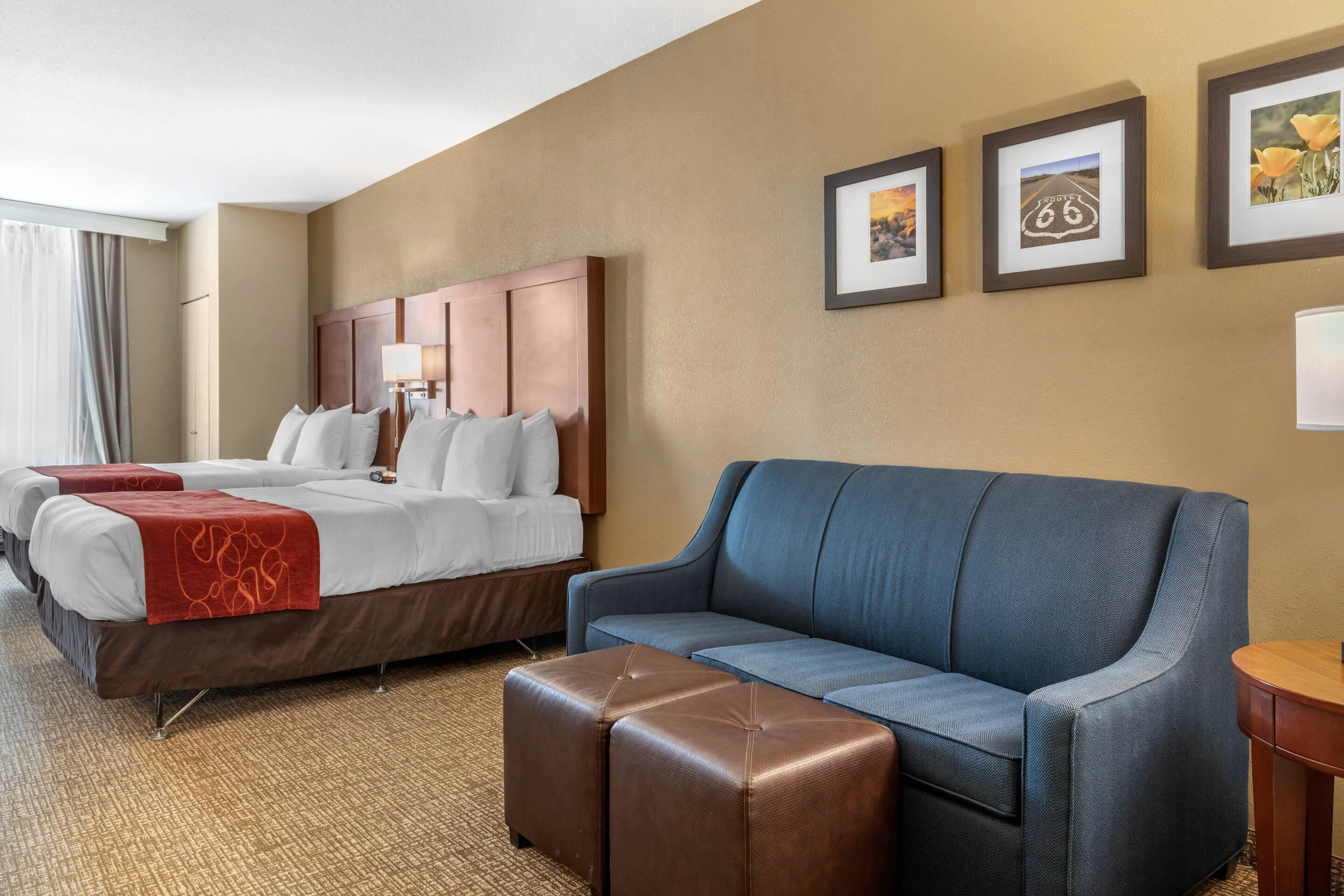 Quarto de hotel com uma cama grande, um sofá azul e um banquinho marrom. Três quadros emoldurados na parede.