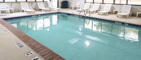 An indoor swimming pool with clear water, marked depth lines, and white lounge chairs arranged around the pool.