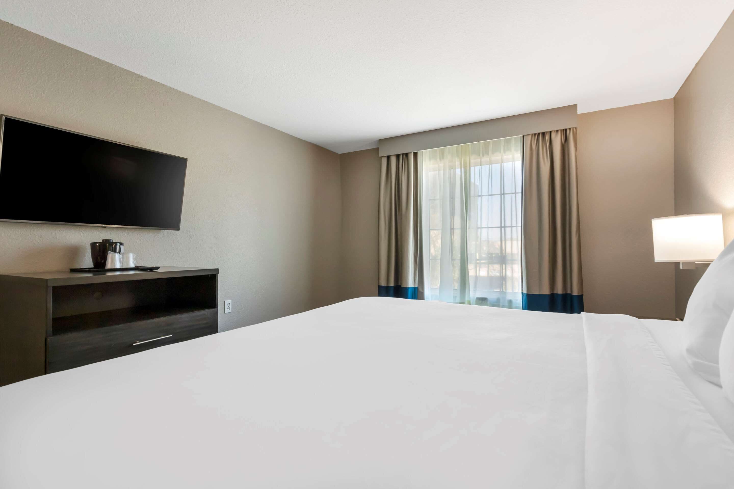 A hotel room with a large bed, a flat-screen TV mounted on the wall, a desk with a lamp, and a window with curtains.