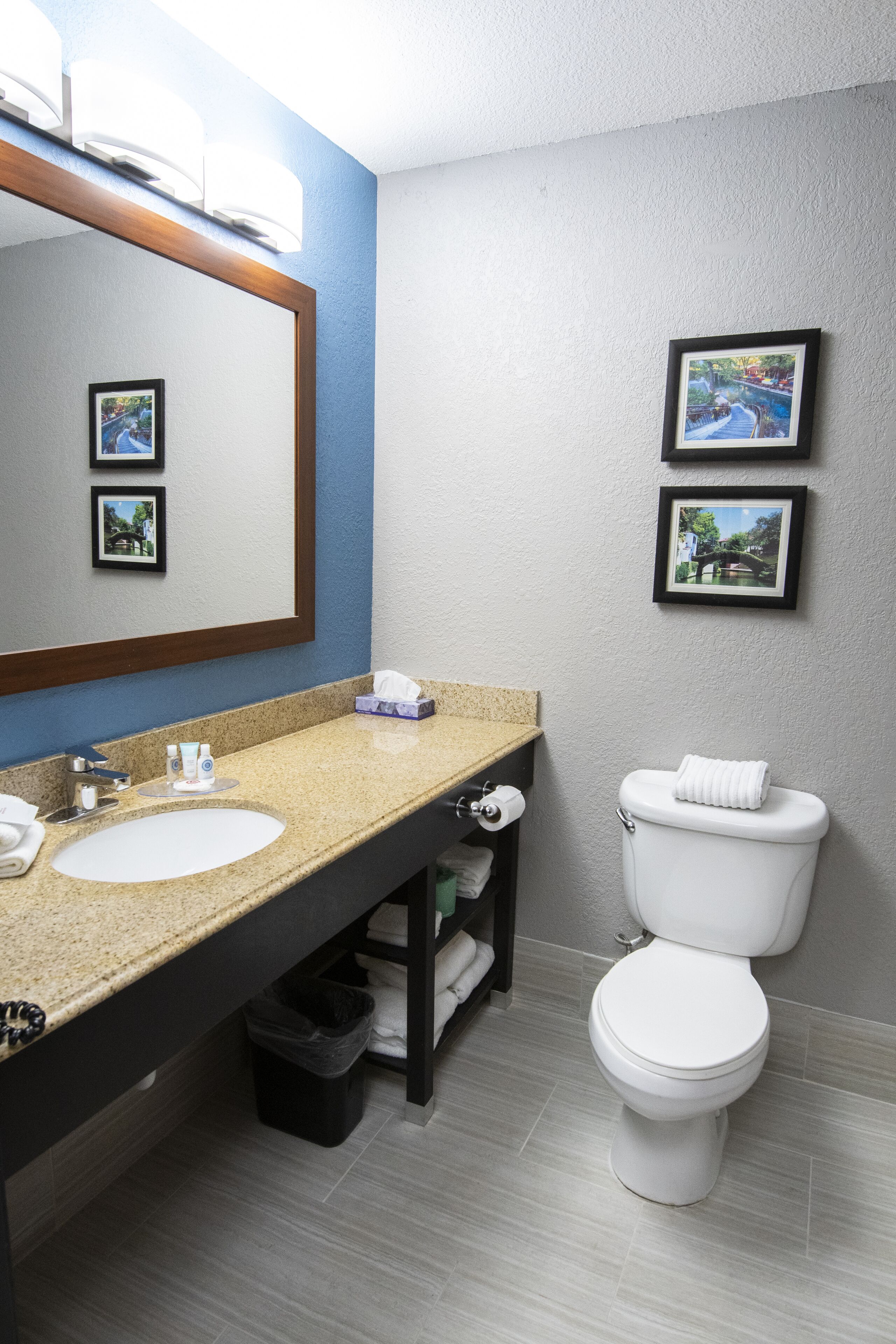 Un baño moderno con lavamanos de granito, un espejo grande, un inodoro y dos cuadros en la pared.