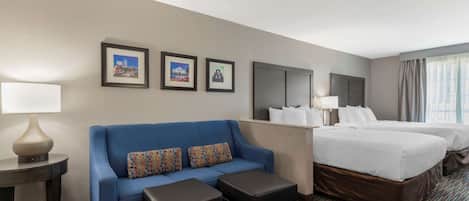 Habitación de hotel con sofá azul, dos camas, una mesita con lámpara y cuadros en la pared.