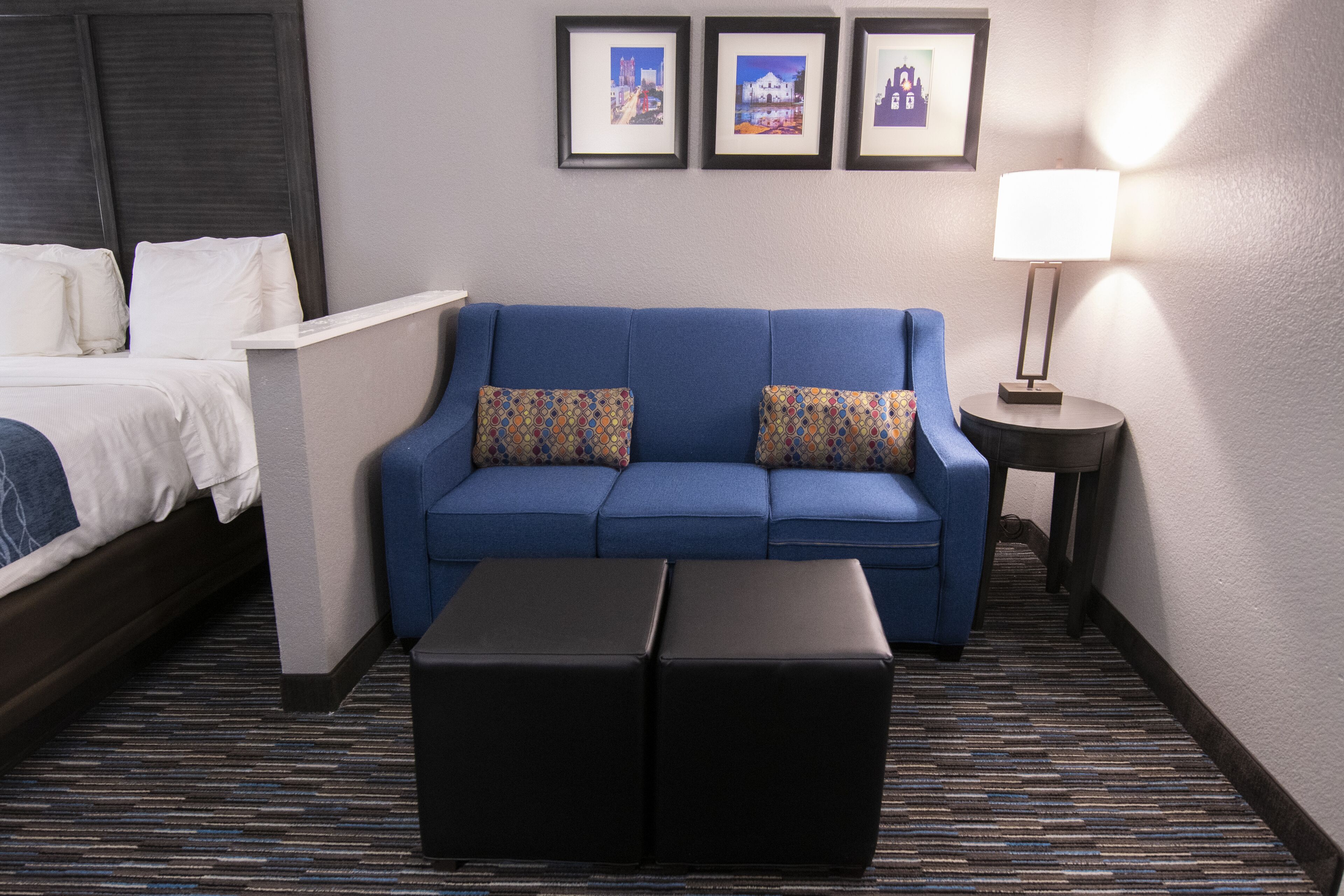 A hotel room with a bed, a blue sofa with patterned cushions, a small table with a lamp, and a carpeted floor.