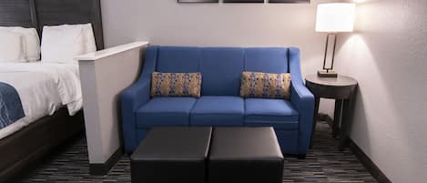 Une chambre d’hôtel avec un lit, un canapé bleu orné de coussins à motifs, une petite table avec une lampe et un sol recouvert d’une moquette.