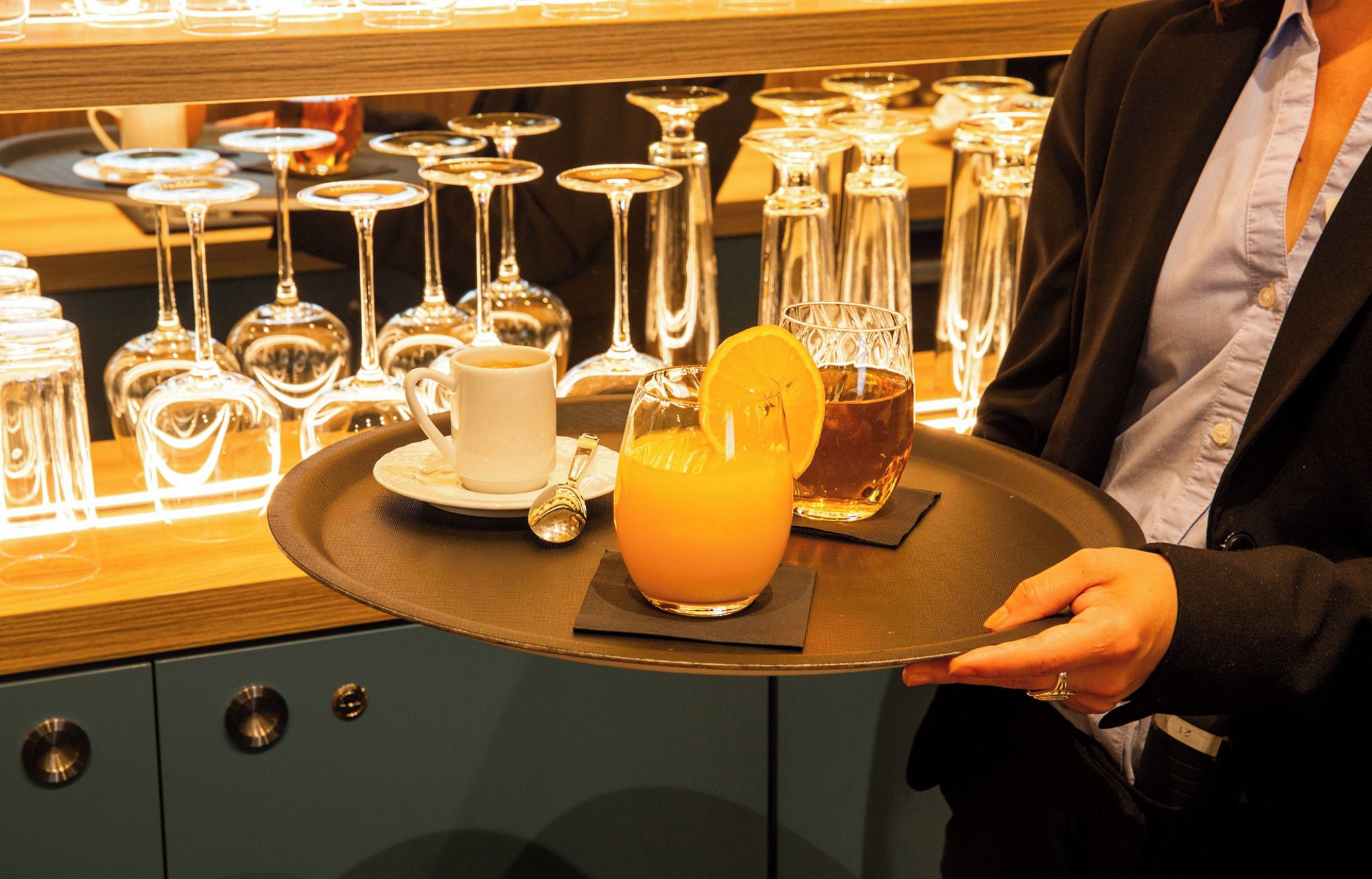 Une personne tenant un plateau sur lequel se trouvent un verre de jus d’orange et une tasse blanche, avec divers verres et bouteilles en arrière-plan.